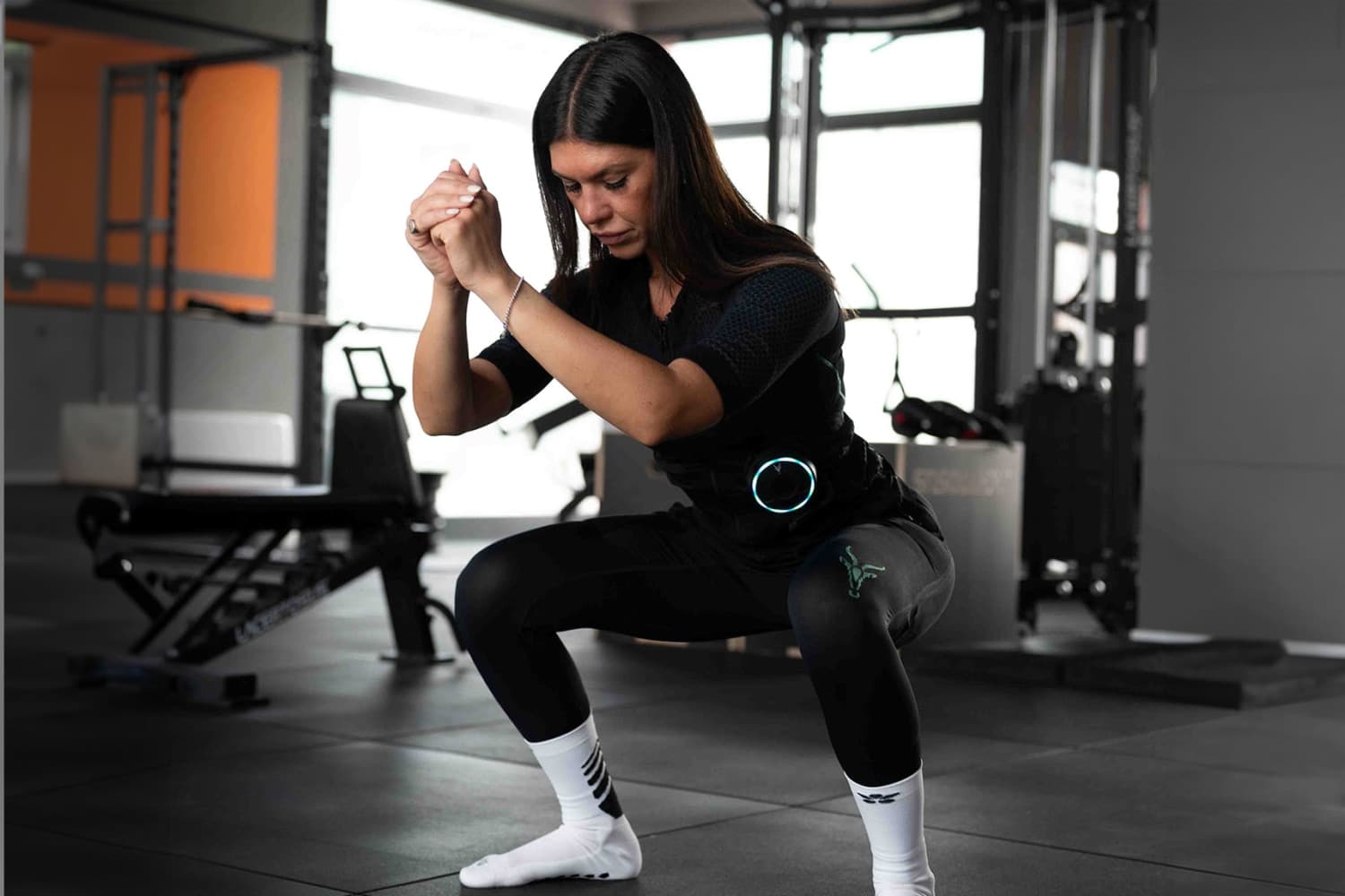 Una giovane donna con la tuta EMS di Antelope esegue uno squat. Si trova in una palestra.