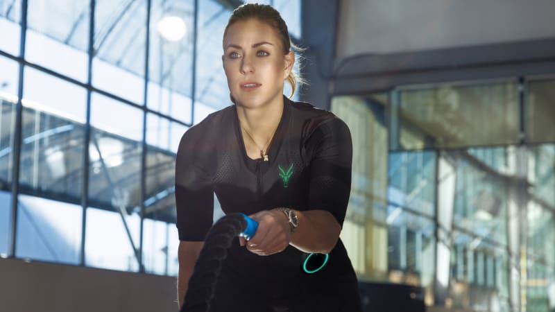 Angelique Kerber integrates training with the mobile EMS suit from Antelope into her workout. She performs a fitness exercise while holding a rope in her hand and looking past the camera on the left. She has tied her blonde hair into a ponytail. The windows of a sports hall can be seen in the background.