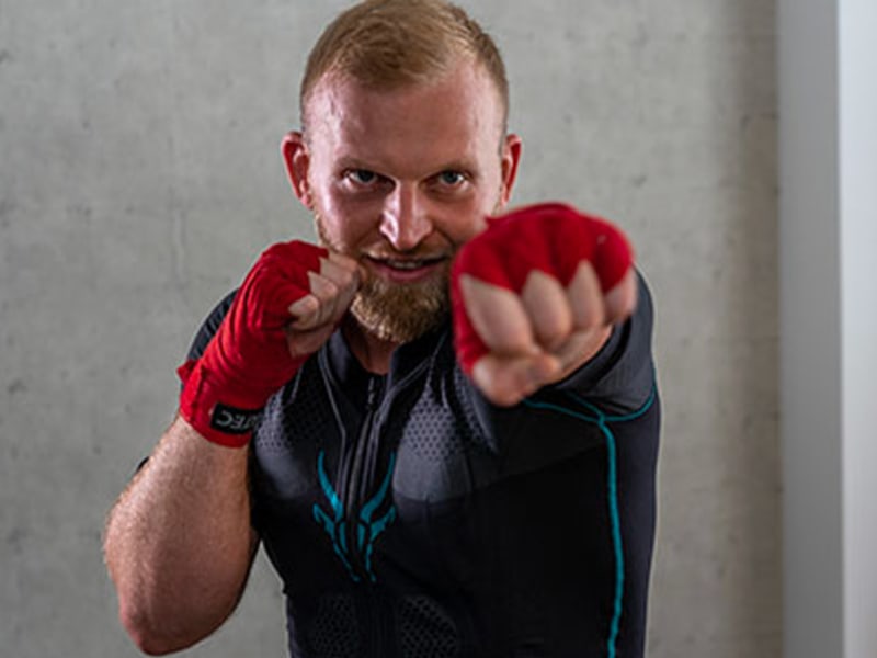 Christoph Rüsseler profite de l'entraînement EMS avec Antelope lorsqu'il fait du kickboxing. Il exécute un mouvement de frappe et regarde la caméra avec concentration. Il porte des bandages de boxe rouges sur les mains. 